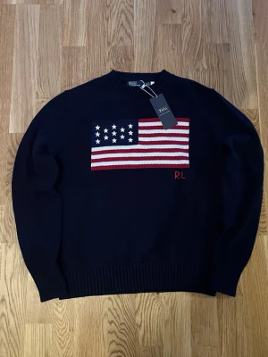 Mörkblå stickad tröja Polo Ralph Lauren - Mörkblå stickad tröja från Polo Ralph Lauren med amerikansk flagga i rött, vitt och blått på bröstet samt RL-broderi. Rund halsringning och ribbade muddar. Perfekt för dig som gillar klassisk preppy stil.