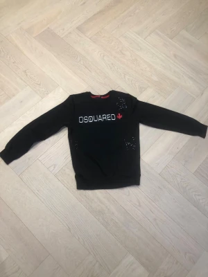 Svart sweatshirt från Dsquared2 - Svart sweatshirt från Dsquared2 med vitt tryck och röd lönnlöv-detalj på bröstet. Tröjan har rund halsringning, långa ärmar och små dekorativa nitar i vitt och rött på framsidan. Perfekt för en trendig och avslappnad stil.