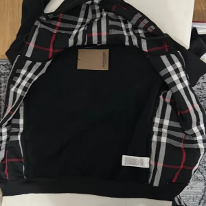 Svart Burberry hoodiejacka med rutig huva - Svart hoodiejacka från Burberry med dragkedja och klassiskt rutig insida i svart, vitt och rött. Jackan har huva med snörning och diskret broderad logga på bröstet. Tillverkad i mjuk bomull, perfekt för en chill och stilren look. Burberry lappen går inte med på köpet.