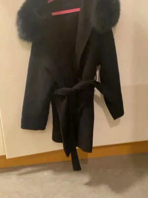 Svart kappa med fuskpäls - TRYCK EJ PÅ KÖP DIREKT!!! Stilren svart kappa med bred krage i fuskpäls och knytskärp i midjan. Kappan har en klassisk siluett och är perfekt för kyliga dagar. Materialet ser ut att vara ullblandning och fuskpälsen ger en lyxig känsla. Passar dig som vill ha en trendig och elegant look.