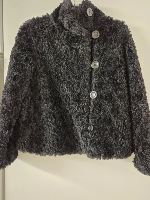 Teddyjacka - Mysig svart teddyjacka med hög krage och stora, silvriga knappar framtill. Jackan har en fluffig och mjuk struktur som ger en cool look. Perfekt för dig som vill ha en varm och trendig jacka till hösten eller vintern.