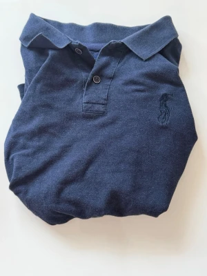 Marinblå pikétröja från Ralph Lauren - Klassisk marinblå pikétröja från U.S polo Assn med korta ärmar och krage. Tröjan har två knappar framtill och den ikoniska broderade loggan på bröstet. Tillverkad i mjuk bomull som känns skön mot huden. Perfekt till jeans eller shorts.