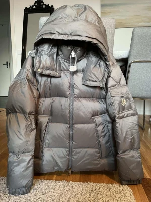 Grå dunjacka från Moncler med huva - Snygg grå dunjacka från Moncler med stor huva och puffig design. Jackan har dragkedja framtill, fickor på sidorna och en ficka med Moncler-logga på ärmen. Tillverkad i glansigt material och perfekt för kalla vinterdagar.