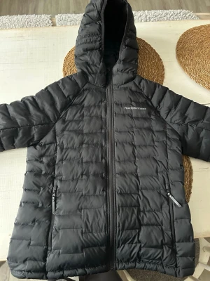 Svart dunjacka Peak Performance - Snygg svart dunjacka från Peak Performance med huva och dragkedja framtill. Jackan har två sidofickor med dragkedja och quiltad design som håller värmen. Perfekt för kalla vinterdagar och har en stilren look som passar till det mesta.