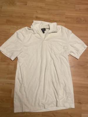 Vit frotté t-shirt från H&M - Vit t-shirt från H&M i mjuk frotté med klassisk krage och bröstficka. Regular fit och korta ärmar gör den enkel att matcha till olika stilar. Perfekt för dig som gillar en clean och chill look.