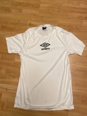Vit sporttröja från Umbro - Snygg vit sporttröja från Umbro med svart logga på bröstet. Tillverkad i ett lätt och snabbtorkande material som passar perfekt till träning eller match. Klassisk rund halsringning och korta ärmar för maximal rörelsefrihet.