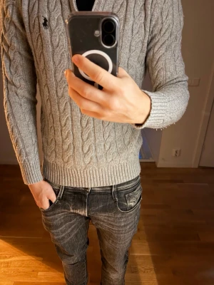 Grå kabelstickad tröja Polo Ralph Lauren - Säljer en grå kabelstickad tröja från Polo Ralph Lauren med klassisk quarter zip och broderad logga på bröstet. Tröjan har ribbade muddar och är tillverkad i mjukt material som passar perfekt till jeans. Otroligt skön! Har av dig vid funderingar!