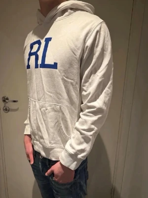 Vit hoodie från Ralph Lauren 🤩🙌 - Snygg vit hoodie från Ralph Lauren med stort blått RL-tryck på bröstet. Hoodien har huva, känguruficka och ribbade muddar vid ärmar och nederkant. Tillverkad i mjukt bomullsmaterial som är skönt att bära.🤩🙌