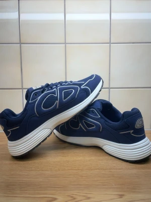Blå Dior sneakers med vit sula - Snygga blå sneakers från Dior med tydlig CD-logga på sidan och sulan. Skorna har en chunky vit sula och detaljer i vitt och grått. Ovandelen är i textil och mesh för en sportig look. Perfekta för dig som vill ha något trendigt och exklusivt.