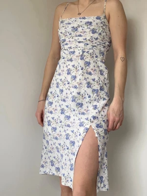 Vit blommig klänning med slits - Söt vit klänning med blått blommönster och smala axelband. Klänningen har en figurnära passform, slits framtill och är tillverkad i ett lätt material som passar perfekt till varma dagar. Storlek: XS