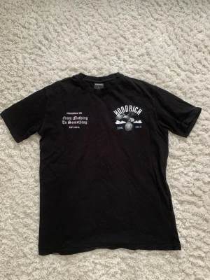 Svart Hoodrich t-shirt med tryck - Svart t-shirt från Hoodrich med tryck på både fram- och baksidan. Framsidan har vit text och logga, baksidan har ett stort motiv med fågel, moln och text. Klassisk passform och rund hals. Perfekt för dig som gillar streetwear.