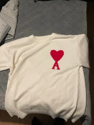 Vit stickad tröja från Ami Paris - Säljer en vit stickad tröja från Ami Paris med röd hjärtlogga och bokstaven A på bröstet. Tröjan har rund halsringning och ribbade muddar. Perfekt för dig som gillar stilrena plagg med en twist.