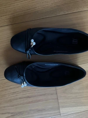 Svarta ballerinaskor med pärldetaljer - Stilrena svarta ballerinaskor från LIX med rund tå och dekorativ rosett framtill. Skorna har fina pärldetaljer och små guldfärgade hängen på rosetten. Tillverkade i ett slätt material som ger en klassisk look.