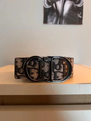 Svartvitt Dior bälte med logga - Snyggt svartvitt bälte från Dior med tydligt CD-spänne i metall. Bältet har ett mönster med Dior-loggor över hela bandet och känns riktigt lyxigt. Perfekt accessoar för att lyfta vilken outfit som helst.