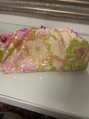 Blommig kuvertväska med paljetter - Kuvertväska i mjukt tyg med broderade blommor i rosa, grönt, orange och vitt. Väskan är dekorerad med små paljetter som ger extra glans. Perfekt för dig som gillar färg och retrostil. Ingen synlig logga eller märke.