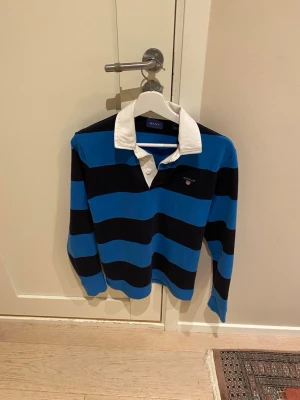 Blårandig rugbytröja från Gant - Klassisk långärmad rugbytröja från Gant med breda blå och svarta ränder samt vit krage. Tröjan har knappar vid halsen och en liten broderad logga på bröstet. Perfekt för en sportig och avslappnad stil.