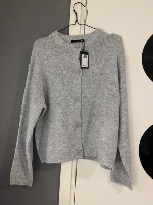 Ljusgrå kofta från lager 157 - Mysig ljusgrå kofta från lager 157 med rund halsringning och stora knappar framtill. Koftan har en rak passform, långa ärmar och är stickad i ett mjukt material som passar perfekt till lager på lager. Enkel och stilren design som är lätt att matcha.