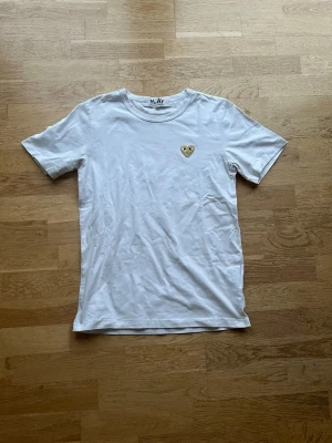 Vit Comme des Garçons Play t-shirt - Säljer en vit t-shirt från Comme des Garçons Play med det ikoniska hjärta-märket i guld på bröstet. Klassisk rund hals och korta ärmar. Perfekt basic med en twist för dig som gillar streetwear och stilrena plagg.