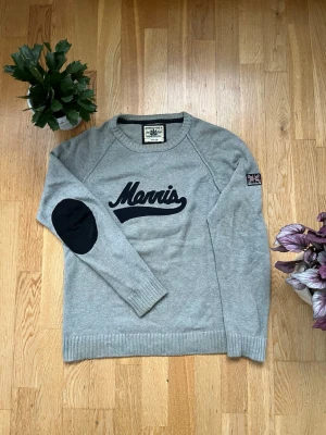 Grå tröja från Morris med patch - Snygg grå tröja från Morris med svart text på bröstet och svart armbågslapp på ena ärmen. Tröjan har rund halsringning, ribbade muddar och en liten brittisk flagga på ärmen. Perfekt för dig som gillar klassisk men sportig stil.