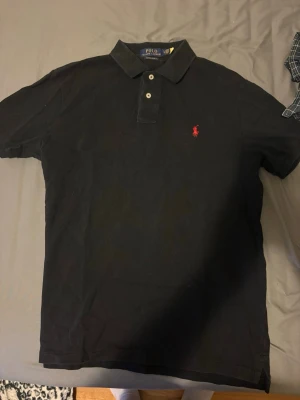 Svart pikétröja från Ralph Lauren - Klassisk svart pikétröja från Ralph Lauren med två knappar och röd broderad logga på bröstet. Tröjan har krage och är kortärmad, perfekt för en clean och stilren look. Materialet är bomull som känns skönt mot huden.