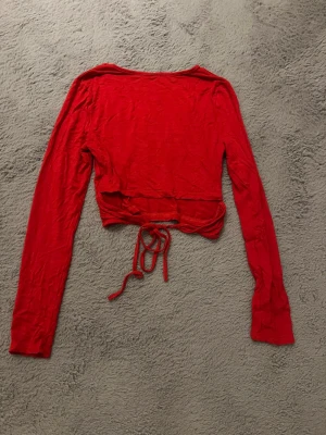 Röd croppad topp med knytning i ryggen - Snygg röd croppad topp med långa ärmar och knyt i midjan. Perfekt för dig som vill ha en enkel men ändå trendig look. Tunn och mjuk i materialet, passar till många olika outfits.