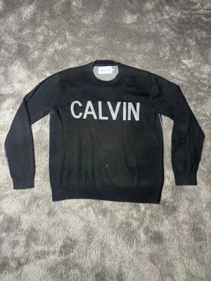 Svart tröja CALVIN Klein Jeans XL - Svart tröja från Calvin Klein Jeans med stor vit text 'CALVIN' på bröstet. Klassisk rund halsringning och ribbade muddar vid ärmslut och nederkant. Perfekt för en clean och stilren look.
