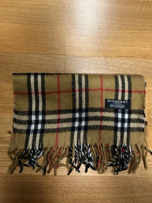 Klassisk beige halsduk från Burberry äkta! - Säljer en ikonisk beige halsduk från Burberry i 100% lammull med det klassiska rutmönstret i svart, vitt och rött. Halsduken har fransar i ändarna och är perfekt för att lyfta vilken outfit som helst. Tveka aldrig på att ställa frågor svarar snabbt!