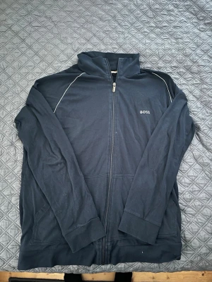 Mörkblå zip hoodie från BOSS - Snygg mörkblå hoodie från BOSS med dragkedja framtill och diskret BOSS-logga på bröstet. Hoodien har vita detaljer längs axlarna och är tillverkad i mjukt bomullsmaterial. Perfekt för en avslappnad och stilren look.