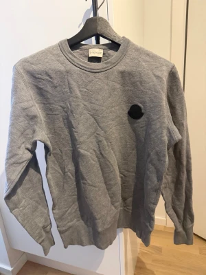 Grå sweatshirt från Moncler - En stilren grå sweatshirt från Moncler med rund halsringning och diskret svart logomärke på bröstet. Tröjan har långa ärmar och ribbade muddar vid ärmslut och nederkant. Perfekt för en avslappnad och trendig look.