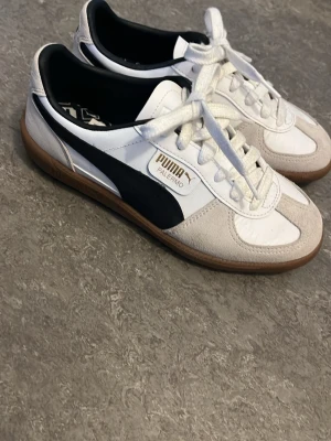 Puma Palermo vita sneakers - Snygga Puma Palermo sneakers i vitt med beige mockadetaljer och svart sidostrip. Klassisk låg modell med gummisula och vita skosnören. Ovandelen är i en mix av läder och mocka, vilket ger en retrovibe. Perfekta för dig som gillar stilrena och sportiga skor. Använd ett fåtal gånger. Säljer pga för små.
