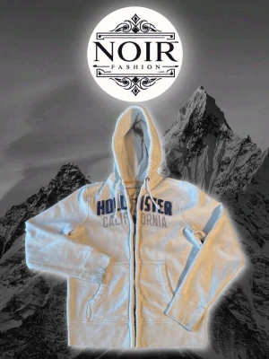 Vit zip-hoodie från Hollister - Snygg vit hoodie med dragkedja från Hollister. Stor tryckt logga i blått och grått på bröstet med texten 'Hollister California'. Utrustad med huva och två fickor framtill. Perfekt för en avslappnad och cool stil.