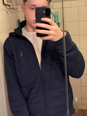 Mörkblå jacka från Ralph Lauren - Mörkblå jacka från Ralph Lauren med dragkedja framtill och klassisk logga på bröstet. Jackan har en quiltad look och hög krage, perfekt för en stilren vardagsoutfit. Passformen är normal och materialet känns mjukt och bekvämt.