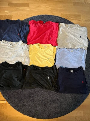 Polo Ralph Lauren t-shirtpaket i olika färger - Säljer ett paket med 9 t-shirts från Polo Ralph Lauren i olika färger: marinblå, röd, grå, gul, ljusblå och svart. Alla har klassisk passform, korta ärmar och den ikoniska broderade loggan på bröstet. Perfekta för en stilren och avslappnad look.