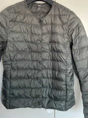 Mörkgrön dunjacka från Uniqlo - Snygg mörkgrön lätt vikt jacka från Uniqlo med rund halsringning och tryckknappar framtill. Västen har ett quiltat mönster och är tillverkad i lätt, glansigt material som ger en clean look. Perfekt för lager-på-lager och enkel att matcha med olika outfits.