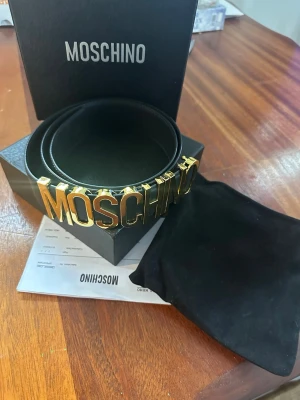 Svart Moschino bälte med gulddetaljer - Snyggt svart bälte från Moschino med stora guldfärgade bokstäver framtill. Bältet är gjort i skinn och har en klassisk, stilren design som verkligen sticker ut. Perfekt accessoar för att lyfta vilken outfit som helst.