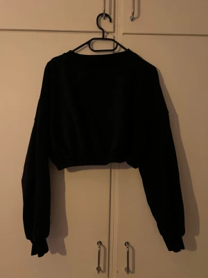 Svart croppad hoodie - Säljer en svart croppad hoodie med långa ärmar och resår i midjan. Hoodien har en enkel och clean design utan tryck eller logga, perfekt till streetwear-stilen. Materialet känns mjukt och bekvämt, troligen bomullsblandning. Passar dig som gillar avslappnad och trendig look.