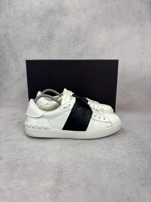 Valentino vita sneakers med svart detalj - Vi har skorna i 39 och 41,5. Snygga vita sneakers från Valentino med en bred svart läderdetalj över mitten och nitar längs hälen. Klassisk låg modell med vita skosnören och diskret logga på plösen. Tillverkade i slätt läder för en clean och modern look.