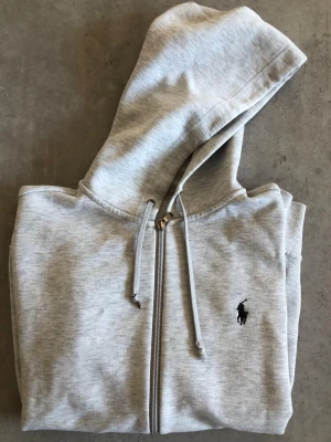 Ljusgrå hoodie från Polo Ralph Lauren - Snygg ljusgrå hoodie med dragkedja från Polo Ralph Lauren. Klassisk design med huva och dragsnören samt den ikoniska svarta logotypen broderad på bröstet. Tillverkad i mjukt sweatshirtmaterial som känns skönt mot huden. Perfekt för en avslappnad och stilren look.