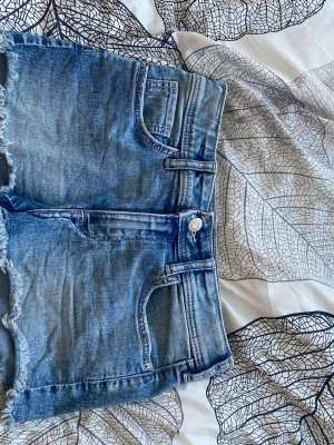 Blå jeansshorts med broderad ficka - Säljer ett par blå jeansshorts med råa kanter och femficksmodell. Ena bakfickan har en stor broderad fjäril som detalj. Shortsen har bälteshällor och klassisk knappgylf. Perfekta för sommardagar!