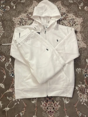 Vitt set från Polo Ralph Lauren - Säljer ett fräscht vitt set från Polo Ralph Lauren med zip-hoodie och matchande byxor. Setet har den klassiska lilla loggan broderad på både tröjan och byxan. Mjukt material och avslappnad passform, perfekt för chill dagar.
