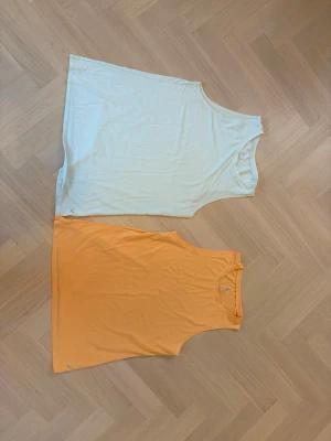 Två ärmlösa linnetoppar med blomtryck - Säljer två snygga linnetoppar i polyester och viskos, en i ljusblå och en i orange. Båda har ett diskret blomtryck framtill och är ärmlösa med rund hals. Perfekta för yoga eller träning. De har ett medel stort tryck på ryggen. De är båda från stadium.