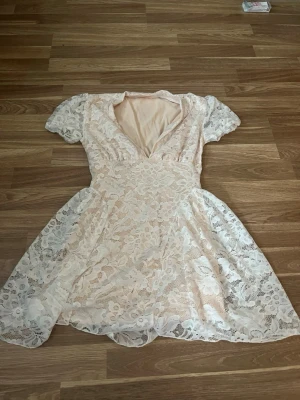 Kort spetsklänning i ljus beige - Söt kort klänning i ljus beige med v-ringning och korta ärmar. Klänningen har ett vackert spetsmönster över hela plagget och en lätt utsvängd kjol. Perfekt för sommarens alla tillfällen!