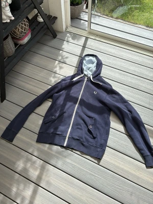 Marinblå hoodie från Fred Perry - Snygg marinblå hoodie från Fred Perry med dragkedja framtill och två fickor med knappstängning. Huvan har ett ljusblått och vitt randigt foder samt vita dragsnören. Perfekt för en avslappnad och sportig stil.