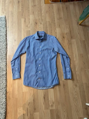 Blå randig skjorta från Eton - Snygg blå skjorta från Eton med tunna vita ränder och klassisk krage. Skjortan har lång ärm, vita knappar och slim fit-passform. Perfekt för dig som gillar stilrena och tidlösa plagg.