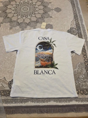 Vit t-shirt från Casablanca - Vit t-shirt från Casablanca med tryck både fram och bak. På ryggen finns ett stort färgglatt motiv med palmer och stadsvy, samt texten 'CASA BLANCA'. 