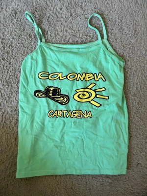 Mintgrön linnetopp med tryck - Mintgrön linnetopp med tunna axelband och tryck i svart och gult med texten 'Colombia Cartagena', en sol och en hatt på framsidan. Perfekt för varma dagar och resor.