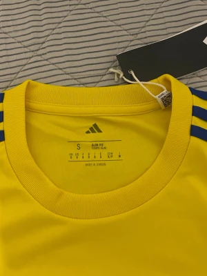 Gul landslagströja Adidas S - Gul t-shirt från Adidas med blå detaljer och tre ränder på axlarna. Tryck med Svenska landslagets fotbollsskola, Stadium, ICA och OBOS på bröstet. Slim fit-modell i funktionsmaterial, perfekt för träning eller match.