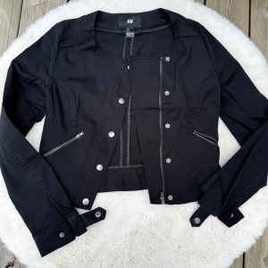 Svart kort jacka från H&M (S) - Snygg svart jacka från H&M i kort modell med rundad krage och silverfärgade dragkedjor. Jackan har dekorativa knappar framtill, två snedställda fickor med dragkedja och spännen vid ärmsluten. Perfekt för en trendig och edgy look.