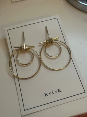 Guldiga dubbla ringörhängen från Hvisk - Säljer ett par trendiga örhängen från Hvisk i guldfärgad metall. Örhängena har två cirklar i olika storlekar som sitter ihop med en cool tvinnad detalj upptill. Perfekta för dig som gillar minimalistisk men ändå unik design.