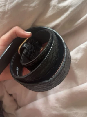 Svart bälte med ormmönster - Snyggt svart bälte med ormmönstrad yta och guldfärgat spänne. Perfekt accessoar för att lyfta vilken outfit som helst. Bältet har en stilren look och passar till både jeans och byxor.